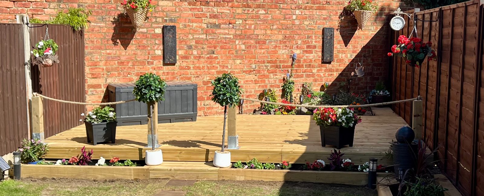 Decked garden with bay trees, planted edges and rope-slung posts against a brick wall