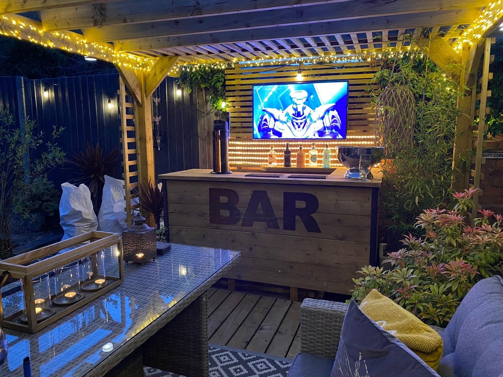 Covered garden bar lit with festoon lights at dusk, planting and seating around it