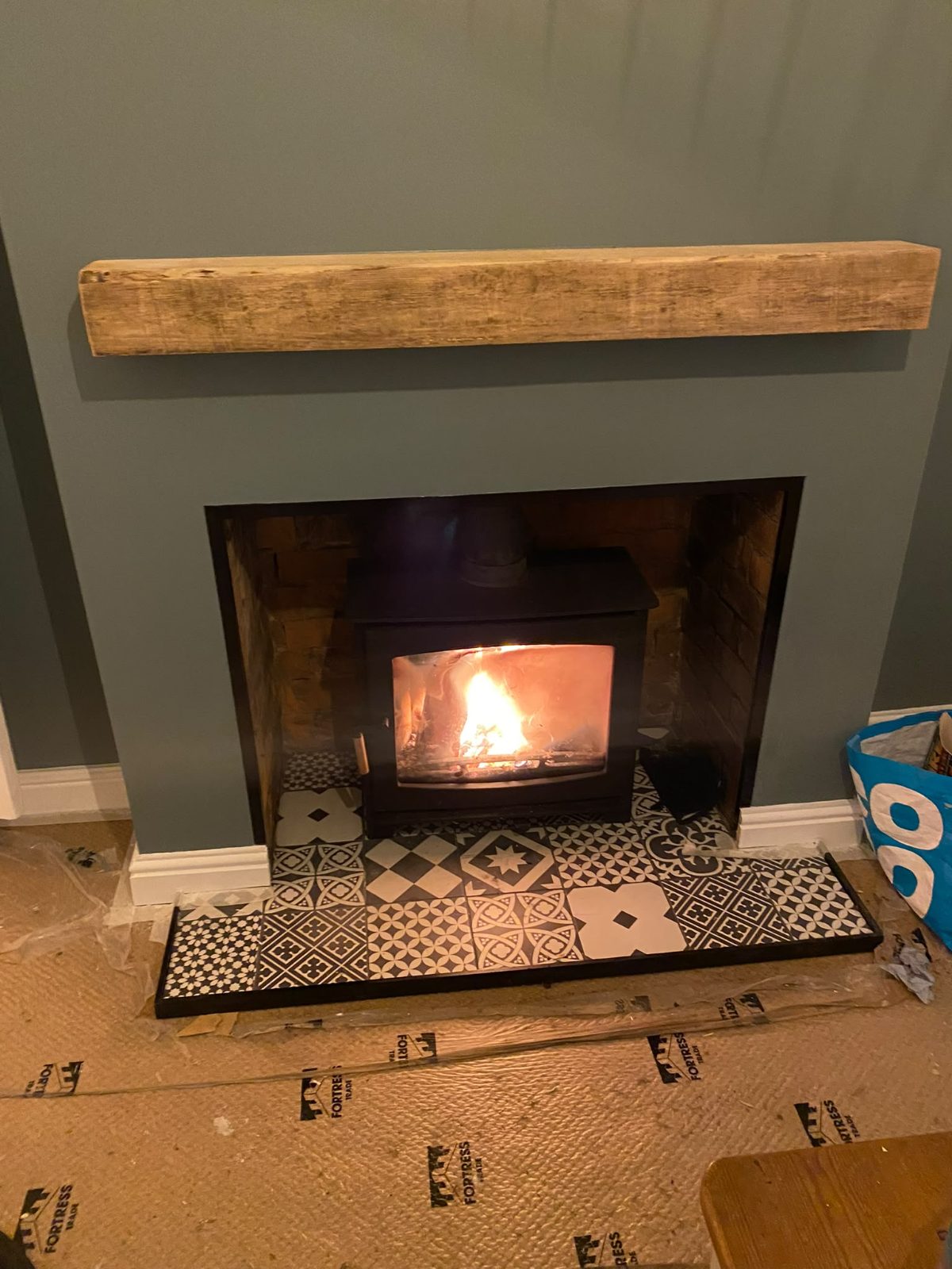Log burner alight in the reopened fireplace under an oak beam mantel, patterned tiled hearth